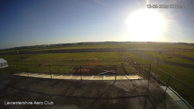 A wide view of a grassy airfield with a runway stretching into the distance under a clear blue sky. The sun is shining brightly in the upper right corner, creating a glare. In the foreground, there is a platform with metal railings and a few structures, possibly equipment or storage areas. The image is timestamped "13-05-2025 07:00:57" in the upper right corner, and the bottom left corner has the text "Leicestershire Aero Club."