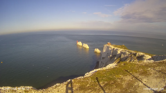 A view from a webcam shows white chalk cliffs extending into the sea, with a grassy top on the right side featuring a small building and a lighthouse. Several large rock formations are visible in the water near the cliffs, and a buoy can be seen floating in the sea. The sky is partly cloudy with patches of blue. A sailboat is visible in the distant right part of the image. The date and time stamp in the bottom left corner indicates it was taken on July 19, 2023, at 8:00:11.