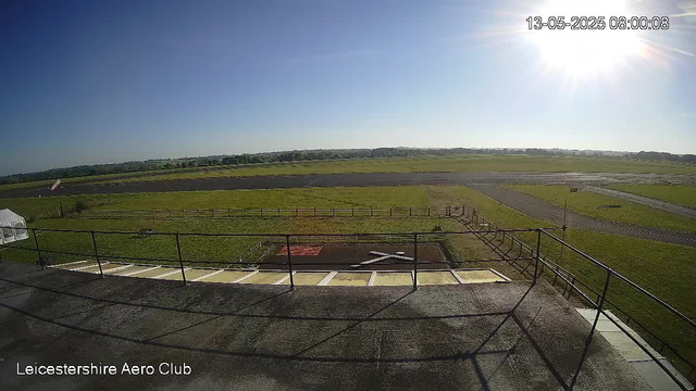 A wide view of an open grassy airfield with a running track, surrounded by fences. The sky is clear and bright with the sun shining from the upper right corner. In the bottom left corner, the text reads "Leicestershire Aero Club," and the top right corner displays a timestamp: "13-05-2025 08:00:08."