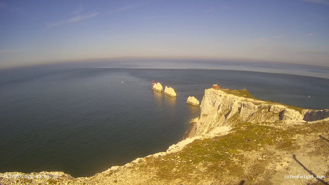 A panoramic view of white chalk cliffs extending into the ocean on the right side, with a small building and antennas on top. Several large rocks are visible in the water near the cliffs, and a red and white lighthouse is situated on one of the rocks. The sky is clear with a few wispy clouds, and the water is calm, reflecting the sky's light.