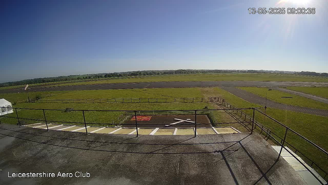 An outdoor view of a small airport runway under a clear blue sky. In the foreground, there is a black railing along a concrete platform with angled parking spaces. Green grass fields extend beyond the platform with a small, red, rectangular patch of ground nearby. A paved taxiway runs parallel to the grassy area, with a few small wind turbines visible in the distance. The timestamp in the upper right corner shows "13-05-2025 09:00:36," and the bottom left corner indicates the location as "Leicestershire Aero Club."