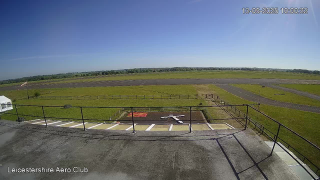 A wide view of an airfield showing a clear blue sky, grassy runways, and taxiways. In the foreground, there is a parking area with designated spaces, a small white tent, and a flagpole. The image is timestamped "13-05-2025 10:00:32" and labeled "Leicestershire Aero Club" in the bottom left corner.