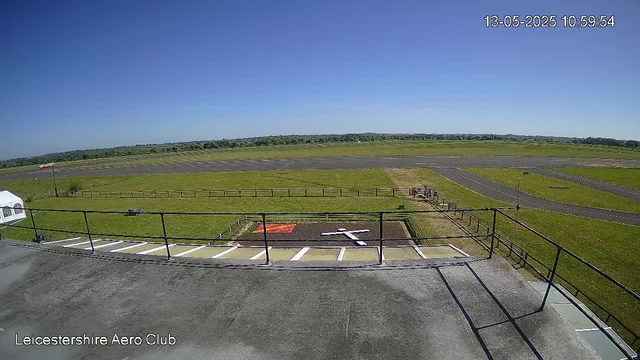 A wide view of an airfield at LeicesterShire Aero Club on a clear, sunny day with a bright blue sky. In the foreground, there is a balcony with a metal railing and parking spaces, one of which has a white vehicle. There is a small fenced area on the grass, with a red and orange object inside, and a couple of people standing near it. The airstrip extends into the distance, surrounded by green fields, with some small structures and trees visible on the horizon. The timestamp in the top right corner shows "13-05-2025 10:59:54."