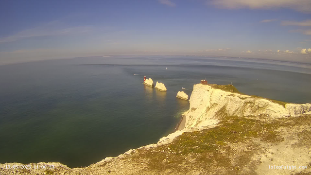A aerial view of white chalk cliffs and rock formations along the coastline, with a small building on the edge of the cliff. The ocean stretches out to the horizon with a few boats visible in the water. The sky is mostly clear with some scattered clouds.
