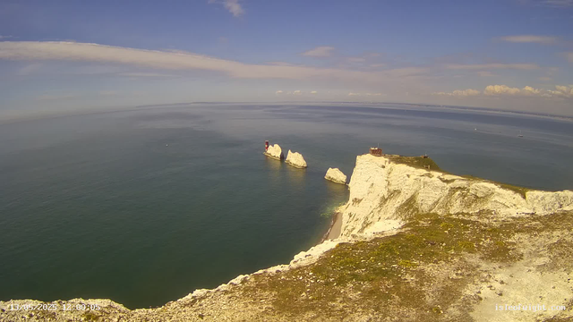 Aerial view of white chalk cliffs overlooking the sea, with several large rocks rising from the water near the base of the cliffs. A red and white lighthouse stands on one of the rocks, and a small building is on top of the highest cliff. The sky is partly cloudy with patches of blue, and the water appears calm. There are a few sailboats visible in the distance. The timestamp on the image reads "13/09/2023 11:00:05".