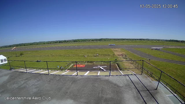 An outdoor view from a webcam shows a clear, sunny day at Leicestershire Aero Club. In the foreground, there is a flat rooftop with a metal railing and parking spaces. Below the rooftop, a small grassy area has a red and yellow object and a couple of people sitting on benches. Beyond that, the grass extends to a runway with two small aircraft parked on the side and a taxiway. In the distance, another aircraft is in motion on the runway. The sky is bright blue with no clouds. The time and date displayed are 13-May-2025 at 12:00:45.