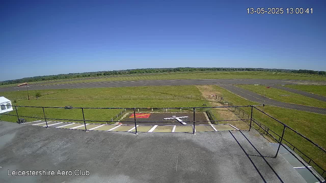 A clear, sunny day at Leicestershire Aero Club with a bright blue sky and no clouds. The image shows a small runway in the distance, surrounded by green fields. In the foreground, there's a rooftop helipad with a white 'H' symbol and a red and yellow marking nearby. A black railing runs along the edge of the rooftop, and a white tent is partially visible to the left. The timestamp in the upper right corner indicates it was taken on May 13, 2025, at 1:00 pm.