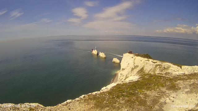 A panoramic view of white chalk cliffs along a coastline, with a small lighthouse and a building on top. The sea is calm, with a few boats visible, and the sky is partly cloudy with streaks of clouds across a blue background. The cliffs extend into the distance, meeting the horizon where the sky and sea converge. The foreground shows the edge of the cliff with some patches of moss or grass.