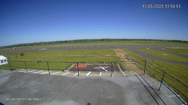 A view from a webcam at Leicesterhire Aero Club showing a clear, sunny day. In the foreground, there is a rooftop with a safety railing bordering the edge, and a designated helipad with a red and yellow marking. Beyond the rooftop, a grassy area surrounds the runway, which stretches across the middle of the image. Two small aircraft are visible on the ground near the runway, with one on the left and another further behind. The sky is bright blue with no clouds, and the horizon line features distant trees and open fields. The date and time in the top right corner read "13-05-2025 13:59:51".