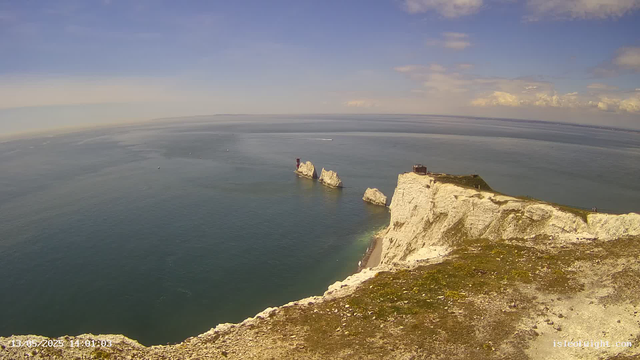 A panoramic view of a coastal landscape featuring white chalk cliffs with patches of green grass on top, extending into the ocean. In the water, several large white rock formations are visible, with a red lighthouse standing on one of the rocks. The sky is mostly clear with some scattered clouds, and the ocean appears calm with gentle waves. A few boats can be seen in the distance. The timestamp in the bottom left corner indicates the photo was taken on May 13, 2025, at 14:01:03.