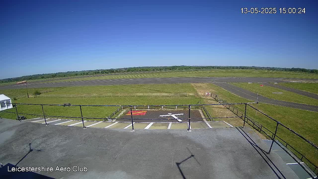A clear day at Leicestershire Aero Club with a view overlooking the runway and surrounding grassy areas. The sky is bright blue with no clouds visible. There are marked parking spaces and a small red and yellow helicopter symbol painted on a section of the ground. The date and time in the top right corner reads 13-05-2025 15:00:24.