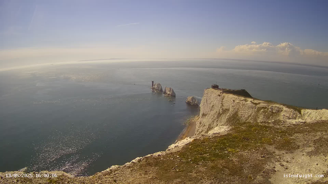 A view of the clifftop at Étretat, overlooking the sea with white chalk cliffs and rock formations in the water. The sky is clear with some clouds on the horizon, and the sunlight reflects off the ocean surface. The image is timestamped with the date and time as May 13, 2025, at 16:00:06.