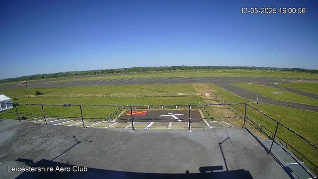 The image shows a clear view of an airfield under a bright blue sky with no clouds. In the foreground, there is a small parking area with marked parking spaces and a white arrow painted on the ground pointing to the right, along with a red rectangular area. The parking lot has black railings along the edge, casting shadows on the surface. To the left, part of a white tent or canopy is visible. Beyond the parking area, there is a grassy field with a yellow wooden fence running horizontally. Further in the background, the airfield’s runway and taxiways are visible, with some small signs and markings. In the upper right corner, the timestamp reads "13-05-2025 16:00:56." The bottom left corner displays the text "Leicestershire Aero Club."