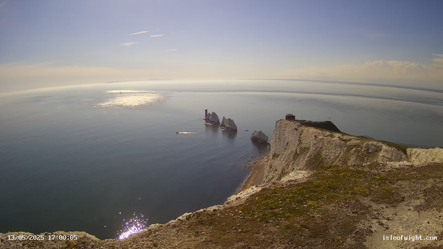 A view from a webcam showing white chalk cliffs overlooking the sea, with a small pathway along the cliff edge. In the distance, there are a few large sea stacks rising from the water, and a lighthouse or small building is situated on the top of the cliffs. The water reflects the sunlight, creating bright spots on the surface, and the sky is mostly clear with a few high clouds. The timestamp indicates the image was captured on May 13, 2025, at 17:00:05.