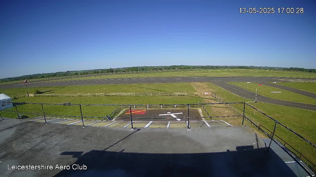 A view of the runway at Leicestershire Aero Club on a clear, sunny day with a bright blue sky. The runway extends into the distance with green grassy areas on either side. There are some taxiways and small vehicles visible in the far background. The foreground shows part of a platform or balcony with black railing, shadowed area, and parking markings. The timestamp in the top right corner reads "13-05-2025 17:00:28."