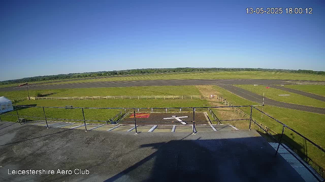 An empty parking area with designated spaces, a helipad marked with a white helicopter symbol, and a black fence surrounding the platform. Beyond the parking lot, there is a grassy field, a small runway, and a clear blue sky with no clouds. In the upper right corner, a timestamp shows the date "13-05-2025" and the time "18:00:12." The bottom left corner reads "Leicestershire Aero Club."
