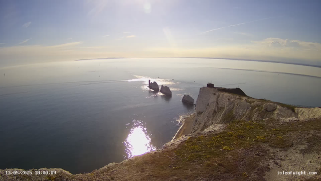 A panoramic view of a coastal landscape with white chalk cliffs and a small building on top. The ocean below reflects the sunlight, with several rock formations and a calm sea extending to the horizon under a clear sky with a few wispy clouds. The scene is illuminated by the sun, casting a bright glare on the water and highlighting the rugged terrain of the cliffs. The timestamp on the bottom left indicates the photo was taken on May 13, 2025, at 6:00 PM.