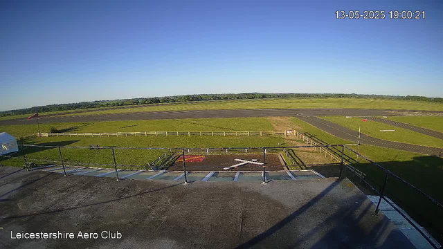 A clear sky over a small airfield with a runway stretching into the distance. The foreground shows a rooftop terrace with a black railing, a flagpole, and a white vehicle parked on the left. There is a red and white sign near the center of the grassy area, which is enclosed by a white fence. The location is the Leicestershire Aero Club, as indicated by text in the bottom left corner. The timestamp in the top right corner reads 13-05-2025 19:00:21.