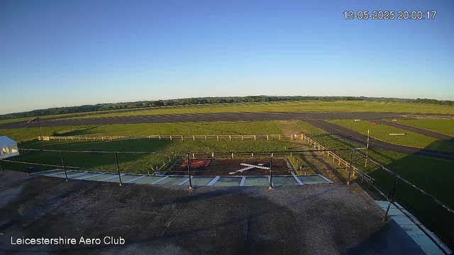An outdoor view from a webcam at Leicestershire Aero Club showing a clear sky, a runway in the distance, a grassy area surrounding the runway, and a small fenced area with a helipad marked by a large white "H" in the foreground. A flagpole with a flag is also visible on the left side of the image. The date and time stamp in the upper right corner reads "13-05-2025 20:00:17."