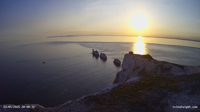 A sunset over a calm ocean with white chalk cliffs in the foreground. Several large rock formations are visible in the water, and a small boat is floating near the cliffs. The sky is mostly clear with a few wispy clouds, and the sun's reflection creates a bright path on the water. The image has a timestamp in the bottom left corner and a website address in the bottom right corner.