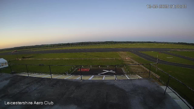 A view from a webcam showing an airfield with a grassy runway and taxiway extending into the distance under a clear sky. In the foreground, there is a concrete platform with a black railing, and a small red and black landing pad with a white arrow painted on it. The bottom left corner has the text "Leicestershire Aero Club." The time and date stamp in the top right corner reads "13-05-2025 21:00:03."
