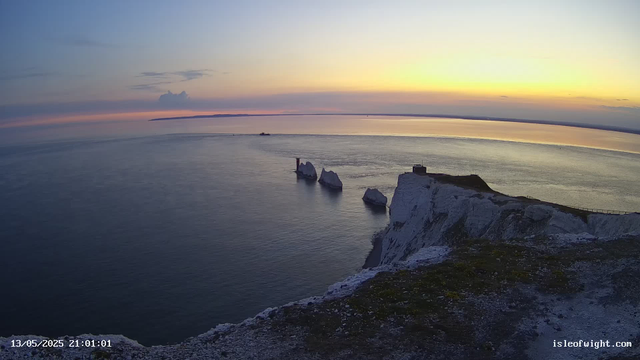 A view of the white chalk cliffs at sunset, with three prominent sea stacks just off the coast. The sky has soft pastel colors with hints of pink, orange, and purple, reflecting on the calm water below. There is a small building or lighthouse on the edge of the cliff, and a path runs along the top. The scene is peaceful and serene, captured during dusk.
