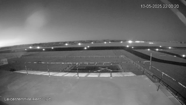 A black-and-white image from a webcam showing an outdoor scene at night with a clear sky and distant horizon. There is a small fenced area and some structures in the foreground, with bright lights illuminating the scene. The timestamp in the top right corner indicates it was taken on May 13, 2025, at 10:00 PM. The bottom left corner displays the text "Leicestershire Aero Club."