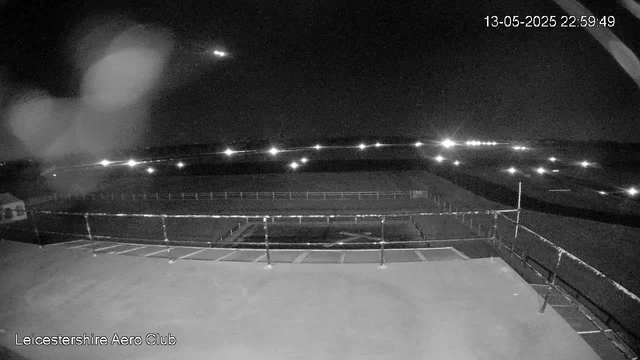A black-and-white nighttime image from a webcam shows an empty outdoor area at Leicestershire Aero Club. The scene includes a fenced circular area, possibly a helipad or sports facility, with distant lights illuminating the horizon. There is a building or structure to the left side of the image. The timestamp in the upper right corner reads "13-05-2025 22:59:49."
