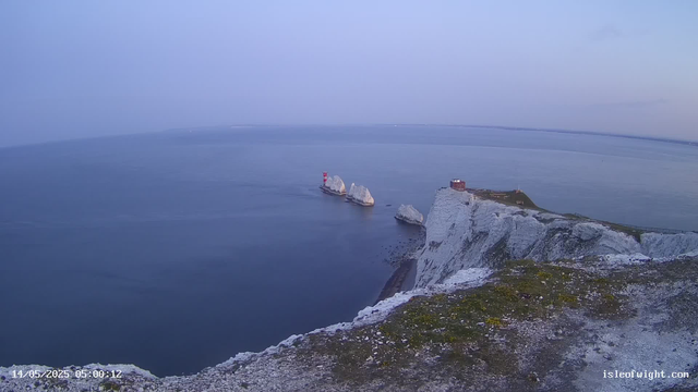 A view of white chalk chalk cliffs with a lighthouse and a small building on top, overlooking the ocean. Large white rock formations are visible in the water near the lighthouse. The sky is clear, and the scene appears to be during early morning or evening.