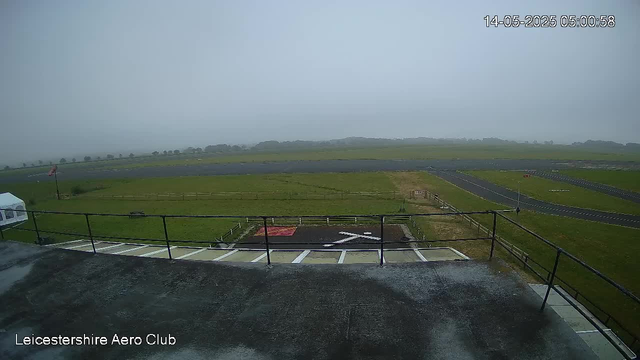 A wide view of an airfield with grassy areas and paved runways under a cloudy sky. In the foreground, there is a balcony or observation deck with a metal railing, and parking spaces are visible below. The image appears to be taken early in the morning or late at night, as indicated by the timestamp in the top right corner. The location is identified as Leicestershire Aero Club.