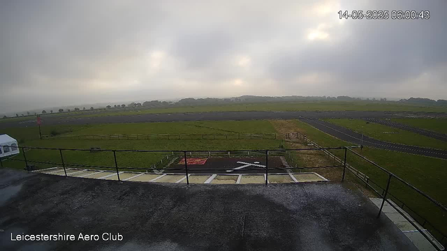 An overcast sky with grey clouds over a flat, grassy landscape at the Leicestershire Aero Club. There is a small helipad with a white helicopter symbol in black, a black border, and a red square on it, located near a fence. The image appears to be taken from a high vantage point, possibly a building terrace or balcony. The timestamp in the top right corner reads "14-05-2025 06:00:43."