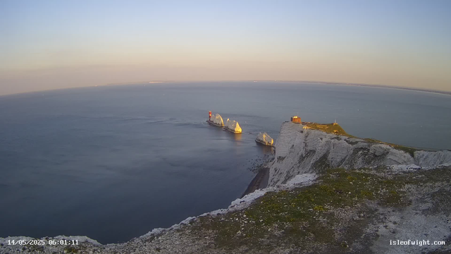 A view of white chalk cliffs overlooking the sea at sunrise. There are several white chalk stacks in the water, with one featuring a red lighthouse. A small building sits atop the highest point of the cliffs. The sky is clear with a pale gradient from light yellow to blue. The date and time are displayed in the lower left corner.