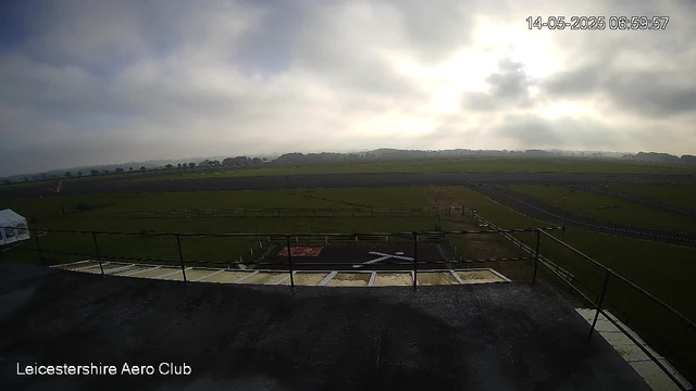 A wide view of an outdoor airport runway and grassy fields under a cloudy sky, with the time and date display showing May 14, 2025, at 6:59 AM. The image is labeled "Leicestershire Aero Club" in the bottom left corner.