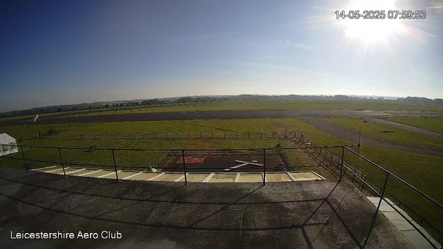 An outdoor view from a webcam at Leicesterhire Aero Club shows a clear, sunny day with a bright sky. In the foreground, there is a rooftop terrace with a metal railing and a few empty parking spaces. Beyond the terrace, there are green grassy fields, a runway or taxiway, and a small winding road. The timestamp in the top right corner indicates it was taken on May 14, 2025, at approximately 8:00 in the morning.