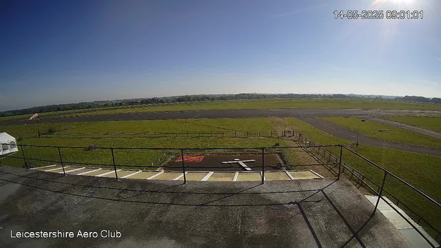 An outdoor view of an airfield taken from a webcam at LeicesterShire Aero Club on a clear, sunny day. The image shows a paved area with parking spaces and a railing in the foreground, a grassy field beyond, and a runway further in the distance. The sky is blue with no visible clouds. The timestamp on the image is 14-05-2025 at 09:01:01.