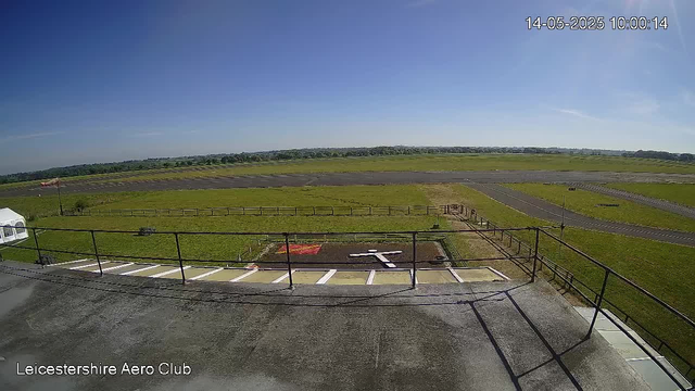 An empty outdoor airfield with a small parking area in the foreground, surrounded by a metal railing. The runway extends into the distance under a clear blue sky. There is a white tent on the left side and a small red and green landing pad visible near the parking area. The image is timestamped "14-05-2025 10:00:14" in the upper right corner, and the bottom left corner displays the text "Leicestershire Aero Club."