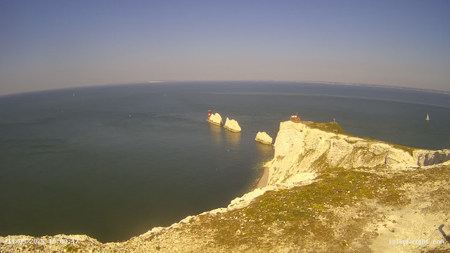 Aerial view of white chalk cliffs along the coastline with a lighthouse and a small building on top. The sea is calm with a few sailboats, and the sky is clear and blue.