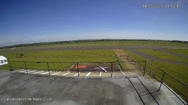 An empty outdoor area at a small airfield with a paved parking area in the foreground, a grassy field, and runways extending into the distance under a clear blue sky. The scene is from a webcam at Leicestershire Aero Club, timestamped May 14, 2025, at 11:00:16.