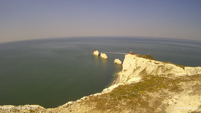 A panoramic view of white chalk cliffs along a coastline extending into the distance, with a small lighthouse and a building perched on top. The ocean surrounds the cliffs, with a few small boats visible on the water, and the sky is clear with a gentle horizon line.