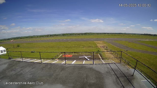 The image shows the view from a webcam at Leicestershire Aero Club, capturing a clear day with blue skies and scattered white clouds. In the foreground, there is a paved area with a safety railing, and a small helicopter landing pad with a red marking and a white helicopter symbol. Beyond, there is a grassy area with a fenced enclosure, along with an airstrip and taxiways stretching into the distance. No people or aircraft are visible in the scene. The timestamp in the upper right corner indicates the date and time: May 14, 2025, at 12:00:12.