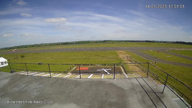 An outdoor view of a large open grassy airfield with a taxiway and some tarmac areas extending into the distance. The sky is mostly clear with a few clouds, and the time stamp in the top right corner reads "14-05-2025 13:01:01." In the lower left corner, there is text that says "Leicestershire Aero Club." The image appears to be taken from a vantage point on a balcony or observation area with a metal railing in the foreground.