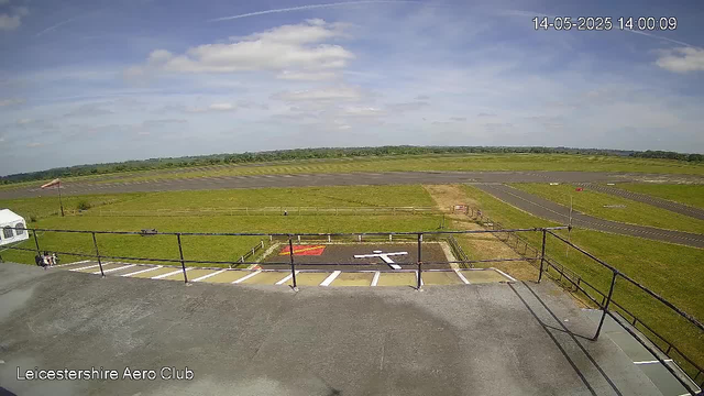 A view from a webcam at Leicestershire Aero Club showing an outdoor area with a runway and grassy fields. The scene includes a small parking area or tarmac section with a railing in the foreground. The sky is partly cloudy with patches of blue, and the timestamp reads "14-05-2025 14:00:09."