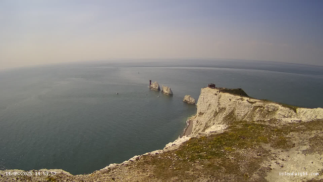 A view from a cliff overlooking the ocean with white chalk formations and a small building or structure near the edge. The water is calm with a few boats visible in the distance. The sky is clear with a soft horizon. The photo appears to be taken from a webcam on a bright, sunny day.