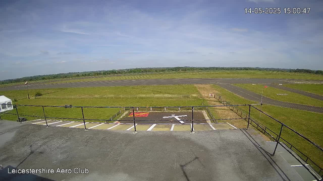 A view from a webcam at Leicestershire Aero Club showing a clear sky with a few clouds, a grassy field, an airstrip with taxiways, and parking spaces with white lines. The timestamp indicates it is 3:00 PM on May 14, 2025. The scene is outdoors, with the horizon stretching into the distance.