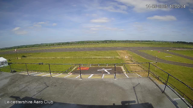 A view from a webcam at Leicester-shire Aero Club showing an empty parking area with marked spaces and a small red and white helipad. Beyond the parking area, there is a grassy field with a taxiway and runway stretching into the distance under a partly cloudy blue sky.