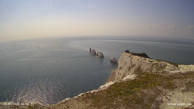 A view from a webcam showing white chalk cliffs meeting the ocean. Several large rock formations are visible in the water near the base of the cliffs. The sky is mostly clear with a few scattered clouds, and the sunlight reflects on the water's surface. The date and time in the bottom left corner are "14/05/2025 16:00:41," and the website "isleofwight.com" is visible in the bottom right corner.