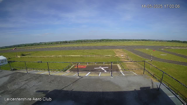 A view of a grassy airfield with a runway and taxiways under a clear blue sky. In the foreground, there is a small parking area with white lines and a directional arrow painted on the ground, along with a red and yellow markings. A black railing surrounds the parking area, and the scene appears to be from a webcam shot taken during daylight hours. The timestamp in the upper right corner shows the date and time as 14-05-2025 at 17:00:06. The bottom left corner contains the text "Leicestershire Aero Club."