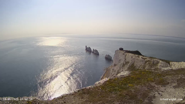 A high-angle view of white chalk cliffs overlooking the ocean during sunset. The cliffs are topped with patches of grass and moss. In the background, the sea reflects the sunlight, creating a shimmering effect. Several large rocks are visible jutting out of the water near the base of the cliffs. The sky is clear with a soft gradient from light to darker tones.