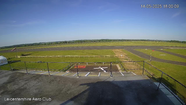 A clear view of an airfield with runways and green grassy areas under a blue sky. There is a fenced area with a small parking space in the foreground, and a windsock on the left side. The image is timestamped 14-05-2025 18:00:09 and labeled "Leicestershire Aero Club" at the bottom left corner.