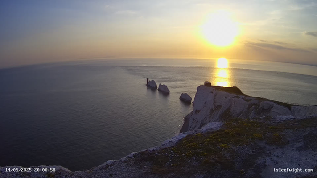 A view of white chalk cliffs overlooking the ocean during sunset, with the sun low on the horizon casting a golden glow. The cliffs have patches of green moss and a small building at the edge. In the distance, several white rock formations rise from the water. The sky is partly cloudy with streaks of clouds illuminated by the setting sun. The timestamp shows "14/05/2025 20:00:58" and the website "isleofwight.com" is visible in the bottom right corner.